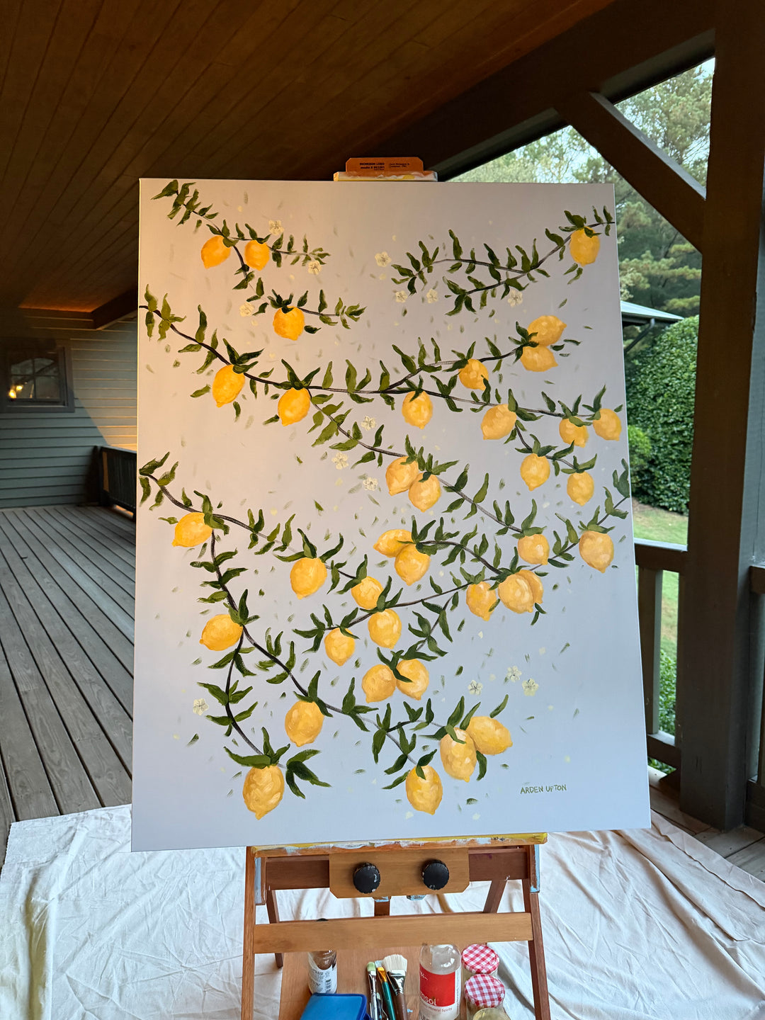 Original Painting by Southern Artist Arden Upton of Lemon tree branches bursting with lemons and green leaves on a light blue background. It is on an easel outside on the porch at Windwood Equestrian. It Would be perfect for kitchens, dining areas, and other places in your home. 