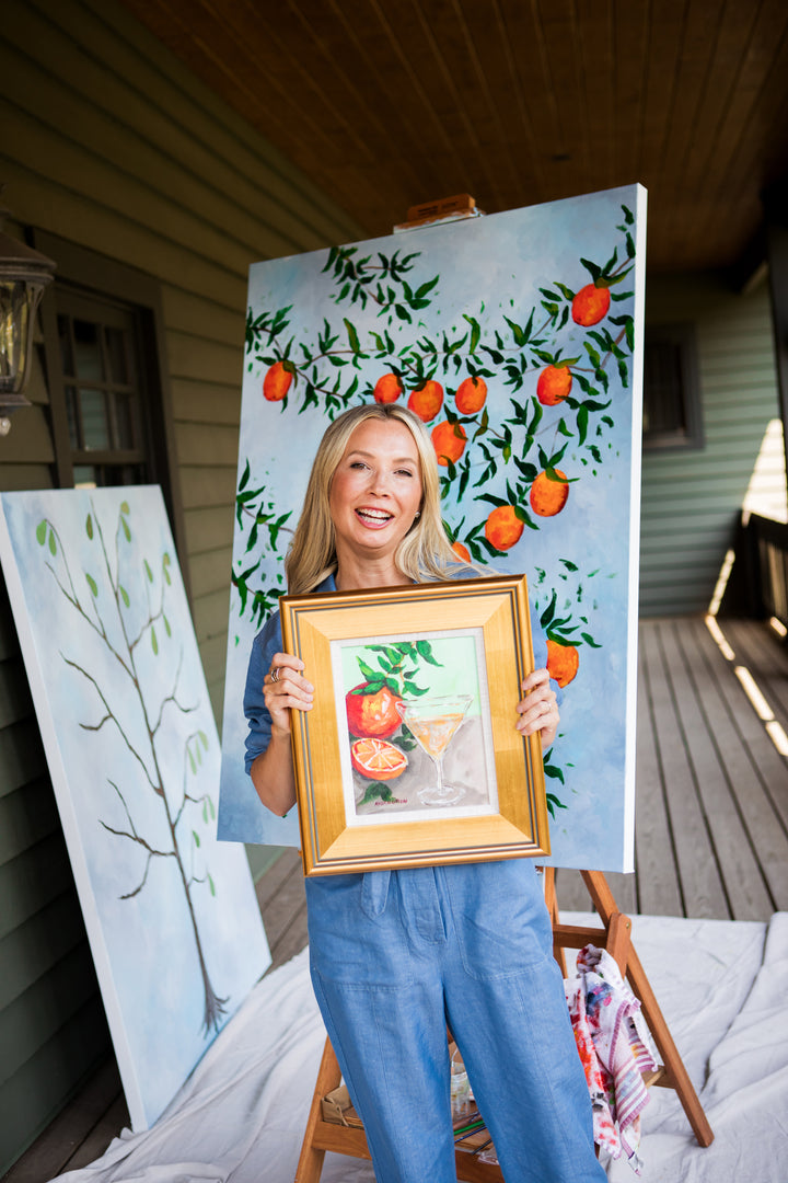 Southern Artist Arden Upton holding a framed painting of oranges and an orange martini framed in a gold frame in front of an easel with a large painting of an orange tree branch.and a pear tree painting in the background. These pieces are great for gathering spaces and dining rooms. 