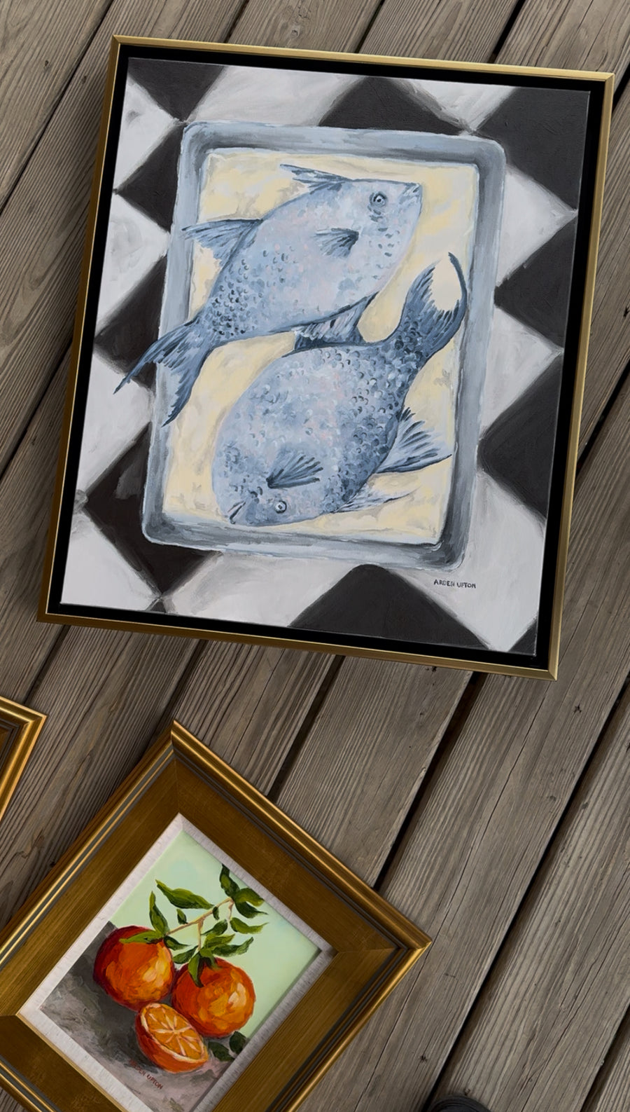 An up close photo of Southern Artist Arden Upton's Blue Fish Oil painting of 2 blue fish on a pan on a black and white checkered table cloth and one of her orange paintings 2 and a half oranges on a grey table with a green background. Both Pieces from her Frank's Table Collection and perfect for Gallery walls, kitchen's dining rooms and brekfast nooks.