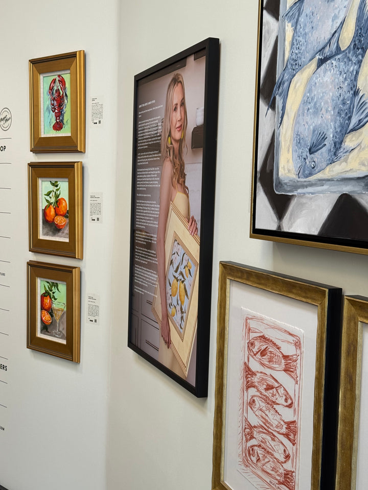 An Angled photo of five pieces of art from Arden Upton's Frank's Table Collection Featuring the lobster painting, two and a half oranges on a grey and green background, and one and a half oranges with an orange martini on a grey and green background. You can also see her two bluefish painting and sketch of five redfish. Perfect for gallery walls and gathering spaces. 