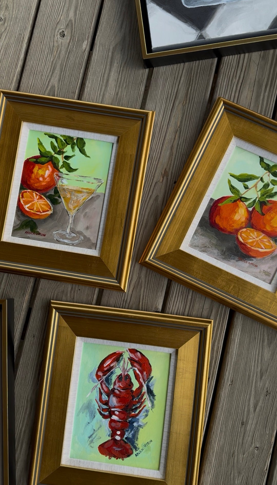 Three framed paintings from Southern artist Arden Upton's Frank's Table Collection of a martini glass, oranges, and a lobster on a wooden surface.