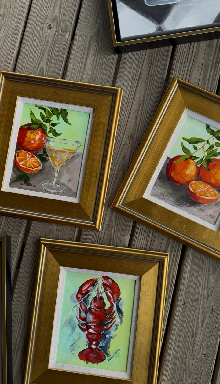 Three framed paintings from Southern artist Arden Upton's Frank's Table Collection of a martini glass, oranges, and a lobster on a wooden surface.