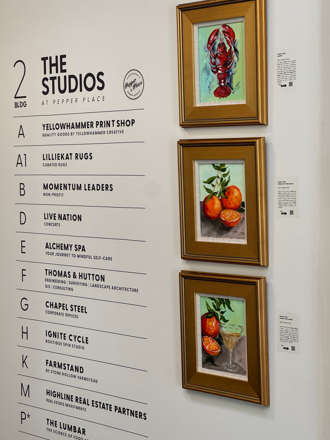 Three gold-framed paintings from Arden Upton's Frank's Table Collection hanging on a white wall in Pepepr Place. The paintings are of a red lobster, oranges with an orange stem and an orange and martini glass. 
