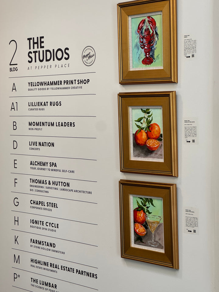 Three gold-framed paintings from Arden Upton's Frank's Table Collection hanging on a white wall in Pepepr Place. The paintings are of a red lobster, oranges with an orange stem and an orange and martini glass. 
