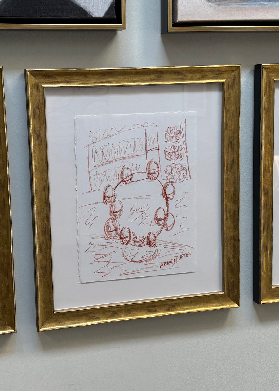 A sketch from Arden Upton's Frank's Table collection depicting farm fresh eggs sitting on the counter top in an egg stand before the ingredients are turned into art. The sketch is matted and framed in a textured gold frame. 
