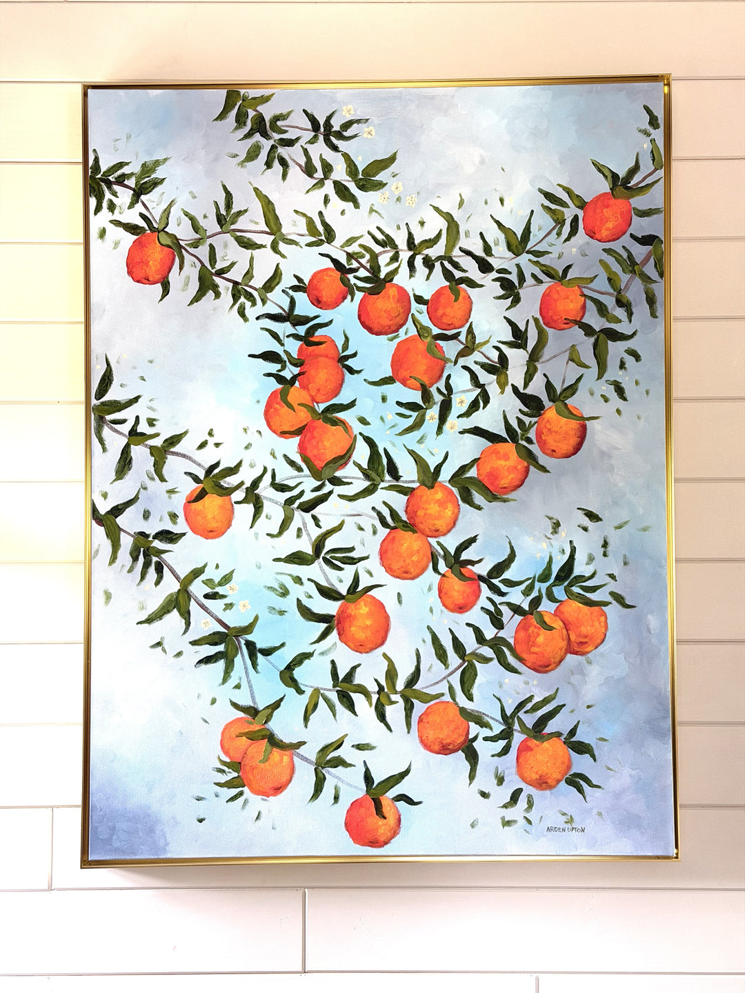 Original oil painting by Southern artist Arden Upton o many branches bursting with ripe oranges and green leaves on a light blue cloudy background. It is hung on a shiplap wall and framed in gold. It would be beautiful in a kitchen or dining room as wlel as many other rooms in a home. 