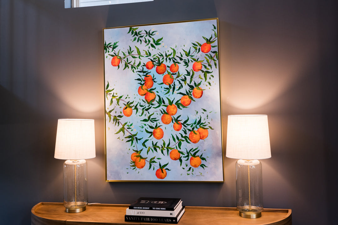 An original painting by Southern Artist Arden Upton of branches full of ripe oranges and green leaves on a light blue background. It is hung on a dark grey wall above a console table with 2 lamps on it. This painting would also look great in a kitchen or dining are to bring happiness into your home.