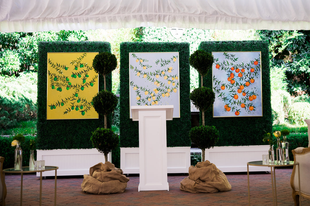 3 original paintings by Southern Artist Arden Upton featured at a luncheon for interior decorators and magazines. The re hung on boxwood walls set behind a white podium. One has green limes on a yellow background, the center painting has yellow lemons on a light blue background and then last one features oranges on a blue background.