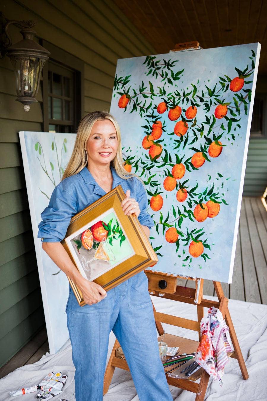 Southern Artist Arden Upton standing in front of her Orangerie V Painting of branches full of ripe oranges and leaves on a light blur background. The pinting on is her Easle on the porch at Windwood with other paintings in the background