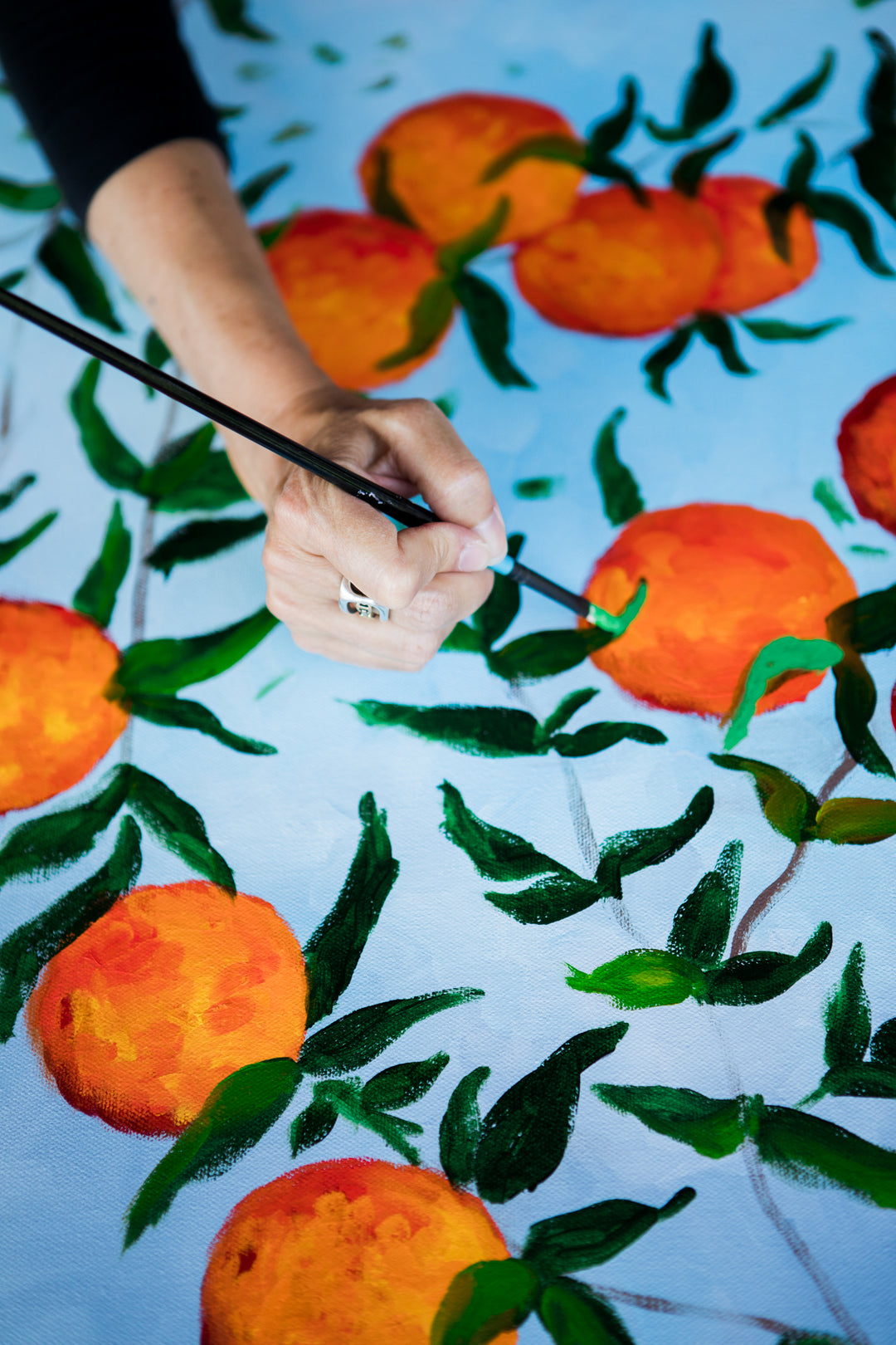 An up close picture of Southern Artist Arden Upton working on the finishing touches of her original oil painting of branches full of ripe oranges and green leaves on a light blue background.
