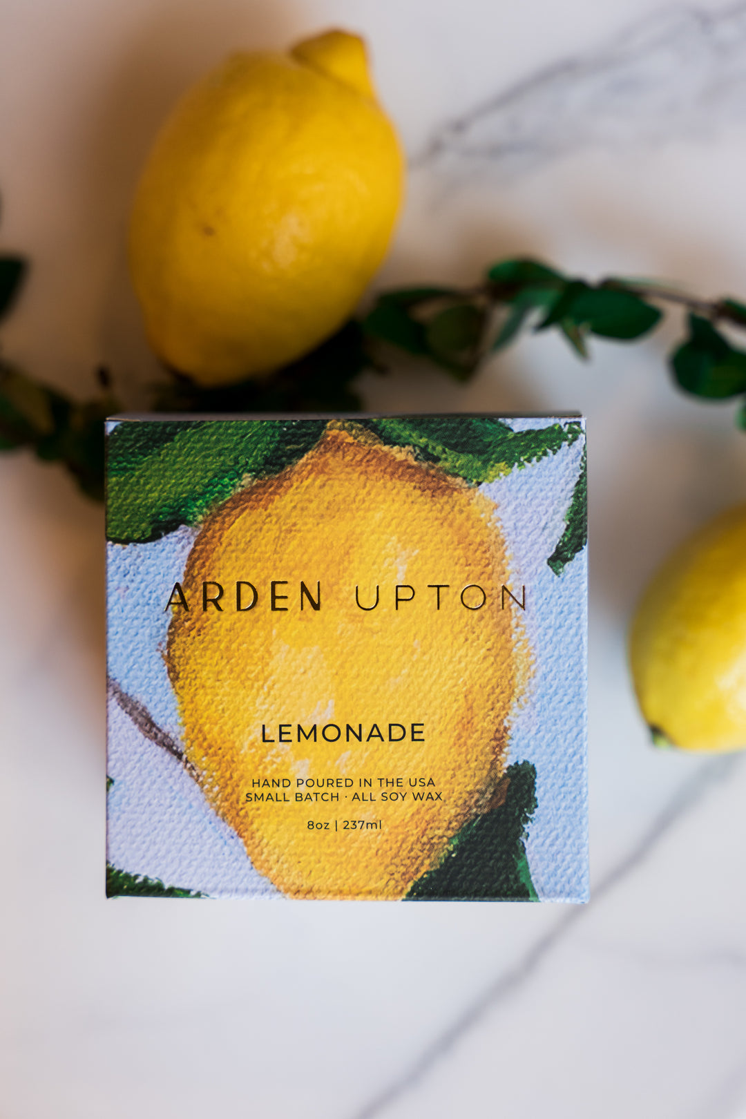 Arden Upton's custom lemonade scented candle in its custom lemon painting with a blue background printed box and gold lettering lying on a counter with greenery and lemons. Makes a great gift and smells wonderful.