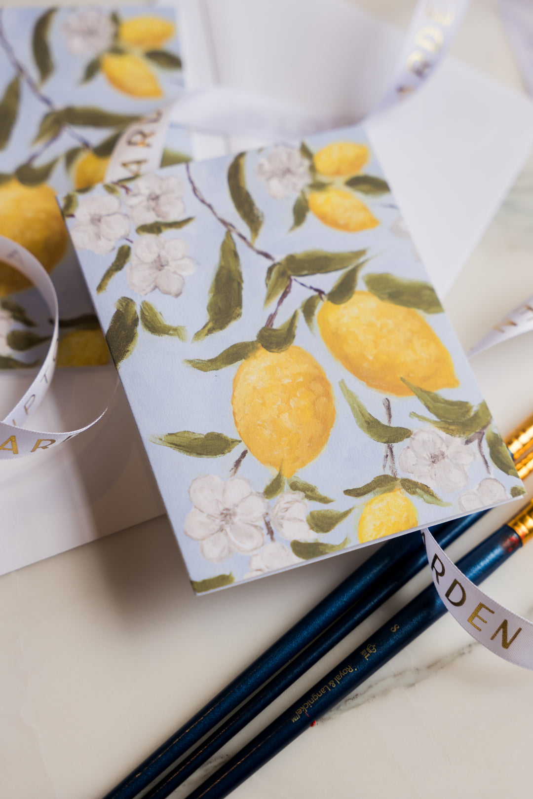 Two folded notecards featuring bold yellow lemons hanging on a branch with soft white flowers and green leaves against a light blue background from Arden Upton's Lemonade Series. The cards are shown with paintbrushes and ribbon and would make a great gift 
