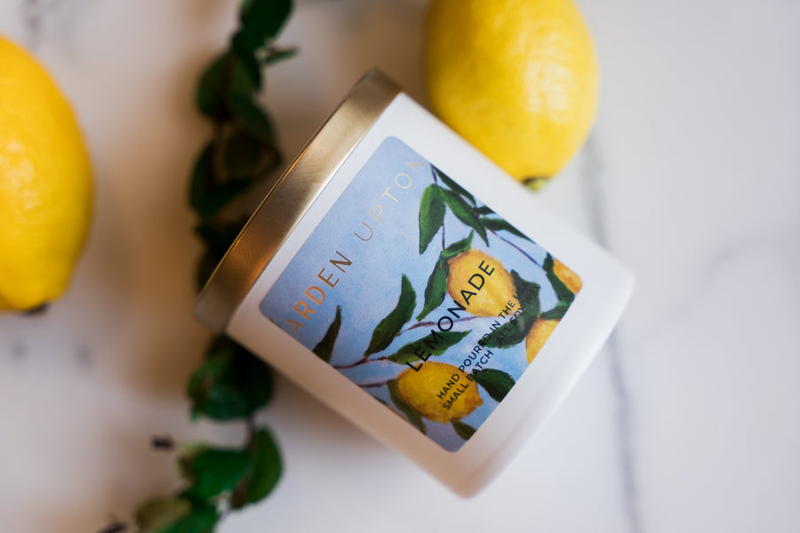 Arden Upton's custom lemonade scented candle in a white fogged glass jar with gold lid and lemon painting print on the label lying on a counter with greenery and lemons. Makes a great gift and smells wonderful.