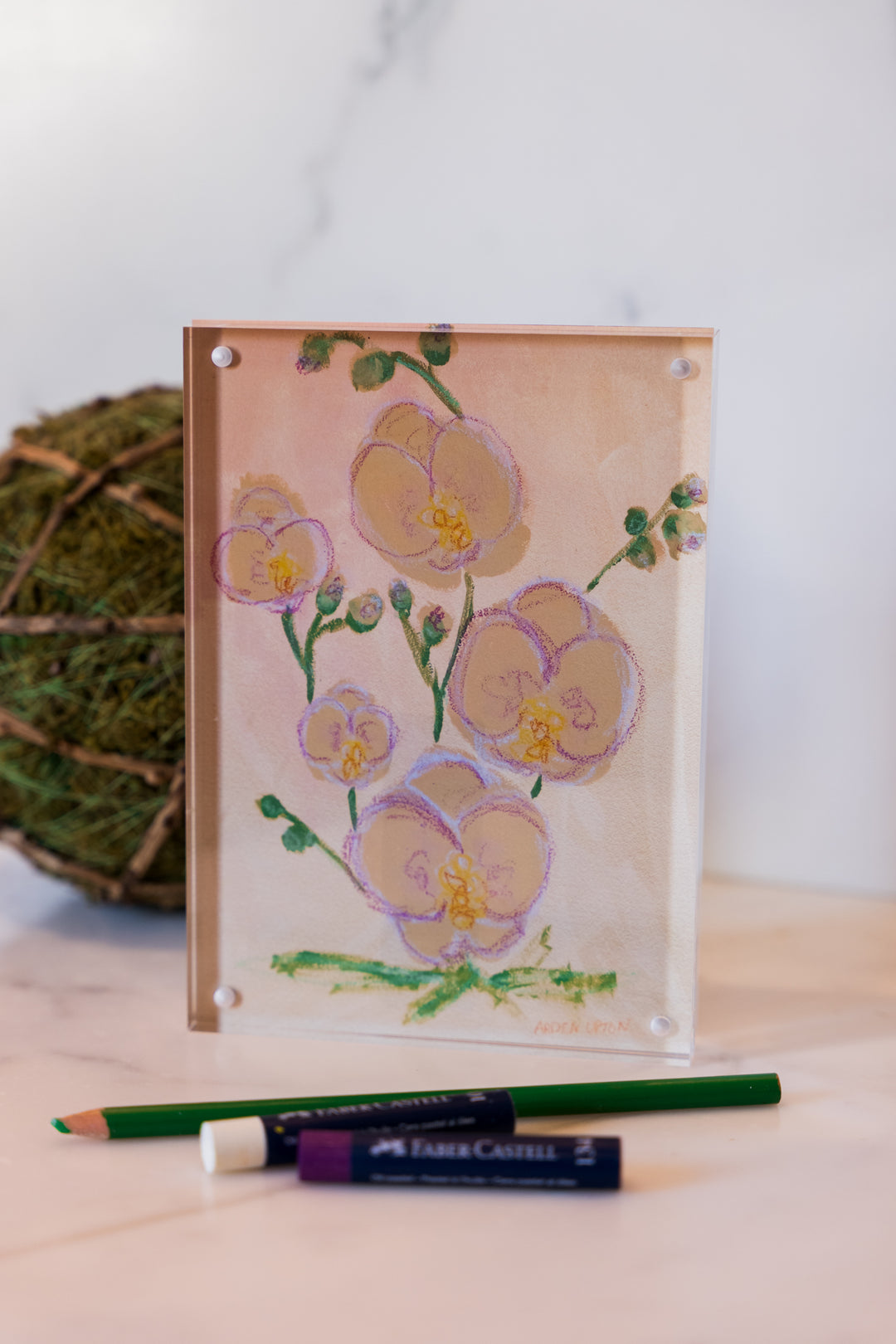A print of a painting of Pink and yellow orchids with green lives from Arden Upton's Flowers that Last Collection  Framed in an acrylic block and staged with art supplies and greenery. These make great teachers gifts and hostess gifts and are beautiful. 
