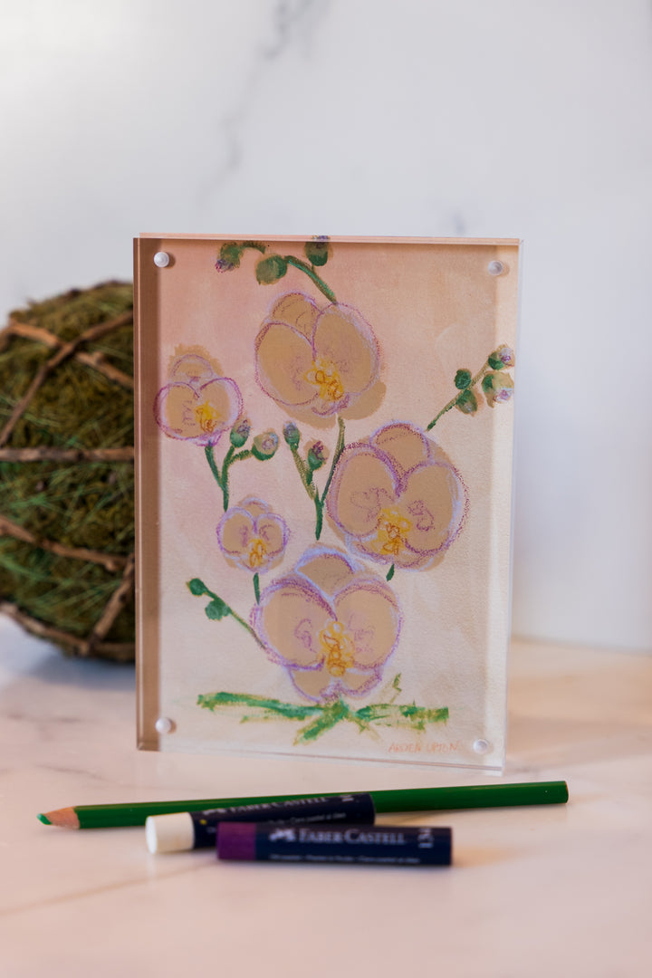 A print of a painting of Pink and yellow orchids with green lives from Arden Upton's Flowers that Last Collection  Framed in an acrylic block and staged with art supplies and greenery. These make great teachers gifts and hostess gifts and are beautiful. 