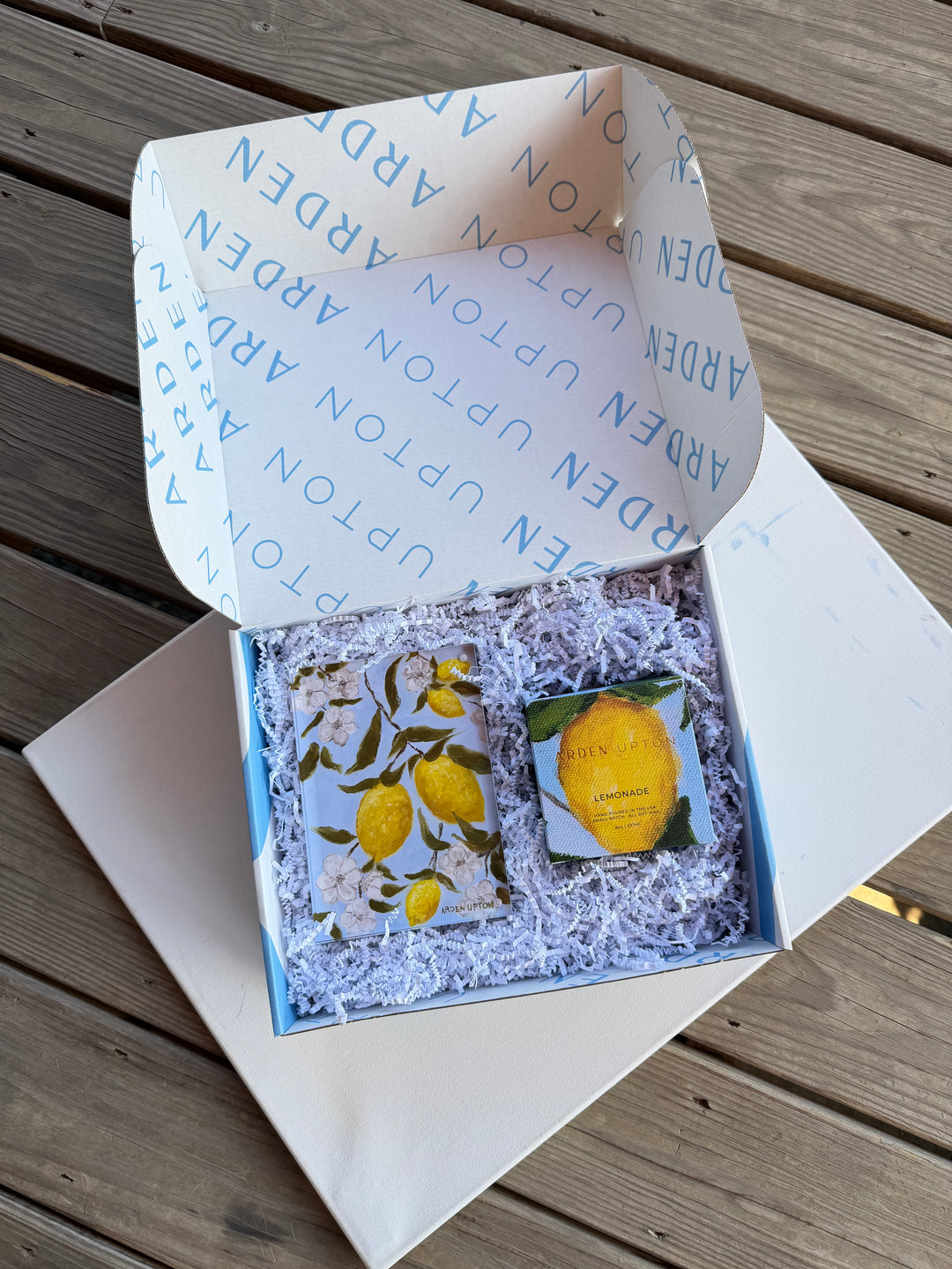 Curated gift set by Arden Upton of her Lemonade print in an acrylic block paired with her signature lemonade scented candle packaged in a box featuring her lemon paintings as well. The gift box is sitting open on a wooden floor and shows Arden's branding on the inside of the box