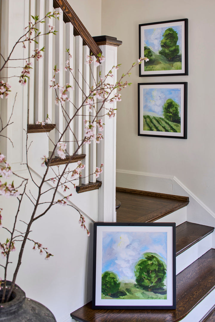 Framed green landscape paintings with blue skies from the Blue Skies Collection by Arden Upton, styled as a gallery wall in a bright entryway. Original artwork and giclée fine art prints perfect for living rooms, hallways, staircases, or gifting