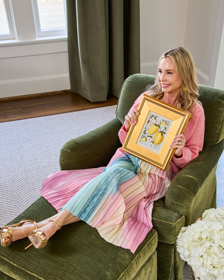 ARDEN UPTON sitting in a green arm chair holding the framed original of one of the pieces from her Lemonade series featuring bright yellow lemons with bright flowers on a beautiful blue background