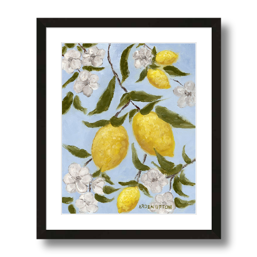 A framed art print featuring a painting from Arden Upton's Lemonade Collection of lemons , leaves and white floral elements against a blue background, with a white mat and black frame.