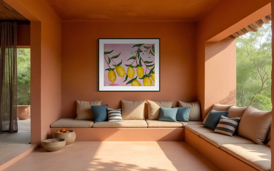 Large scale print of a painting from Arden Upton's Lemonade Series of bright yellow lemons with green leaves and white flowers hanging from a branch on a pink background styled in an outdoor seating area
