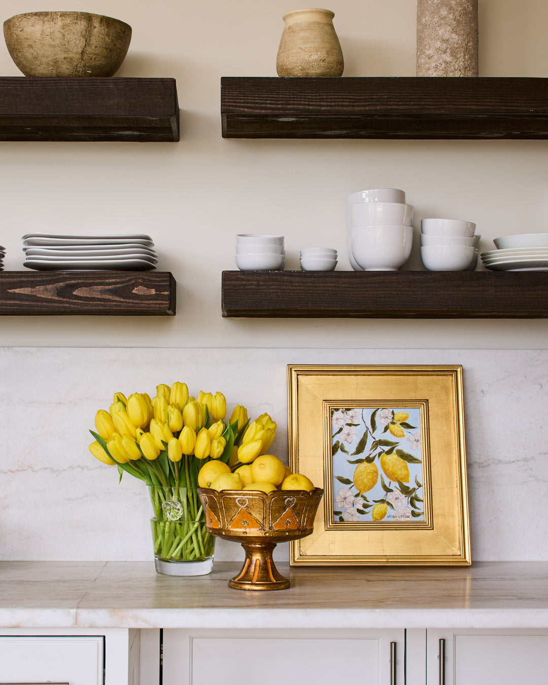 Kitchen counter with a bowl of lemons, yellow tulips and a framed painting from Arden Upton's Lemonade Series featuring bright yellow lemons with white flowers and green leaves on a bright blue background. 