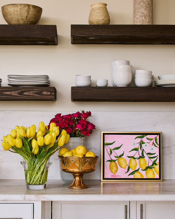 A painting from Arden Upton's Lemonade Series of bright yellow lemons, green leaves and white flowers on a pink background sitting on a kitchen counter with a bowl of lemons, a vase of yellow tulips and a vase of red roses. 