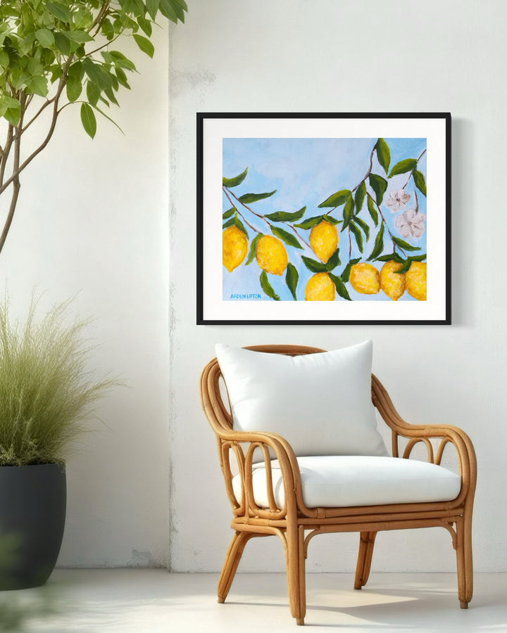 A framed art print featuring a painting from Arden Upton's Lemonade Collection of lemons , leaves and white floral elements against a blue background styled over a chair
