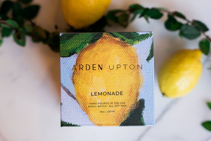 Arden Upton's custom lemonade scented candle in its custom lemon painting with a blue background printed box and gold lettering lying on a counter with greenery and lemons. Makes a great gift and smells wonderful.
