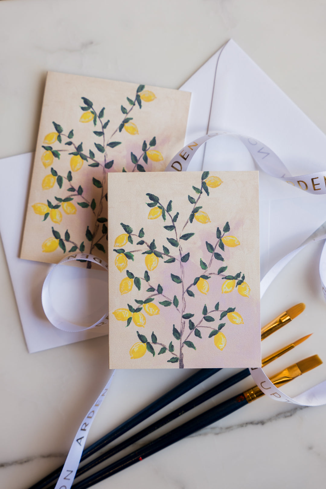 Two folded greeting cards with Arden Upton's lemon tree painting of bright yellow lemons and green leaves on the tree printed on them laying on a marble surface with art supplies. These Notecards make a great gift.