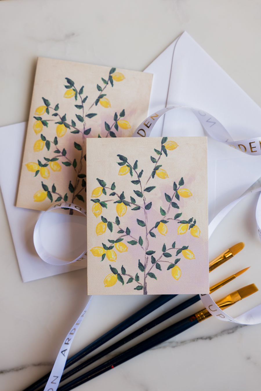 Two folded greeting cards with Arden Upton's lemon tree painting of bright yellow lemons and green leaves on the tree printed on them laying on a marble surface with art supplies. These Notecards make a great gift.