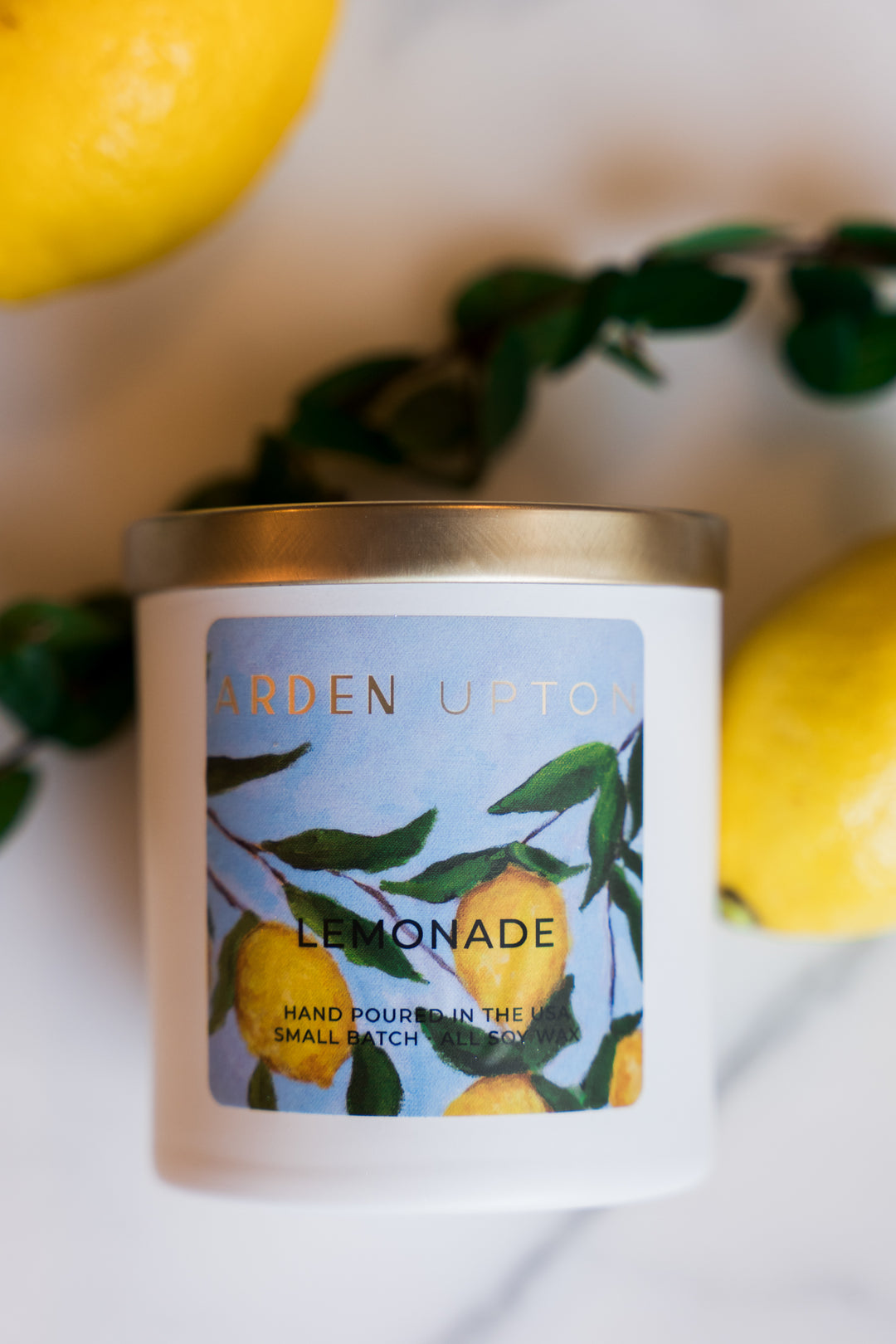 Arden Upton's custom lemonade scented candle in a white fogged glass jar with gold lid and lemon painting print on the label lying on a counter with greenery and lemons. Makes a great gift and smells wonderful.