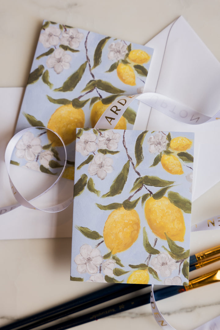 Two folded notecards featuring bold yellow lemons hanging on a branch with soft white flowers and green leaves against a light blue background from Arden Upton's Lemonade Series. The cards are shown with paintbrushes and ribbon and would make a great gift 