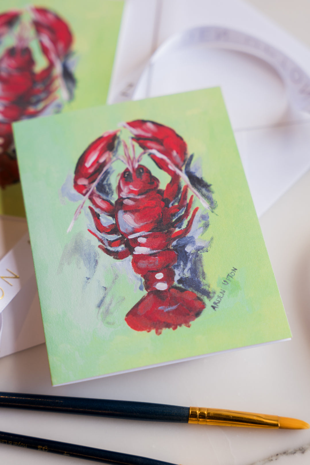 A photo of a note card featuring Arden Upton's original lobster painting of a bright red, whimsical lobster on a green background. The note cards come in sets of 6 and are a great gift idea. In the photo, the note cards are surrounded by paintbrushes. 