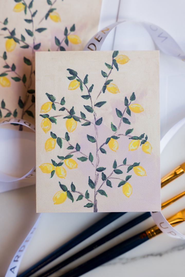 Two folded greeting cards with Arden Upton's lemon tree painting of bright yellow lemons and green leaves on the tree printed on them laying on a marble surface with art supplies. These Notecards make a great gift.