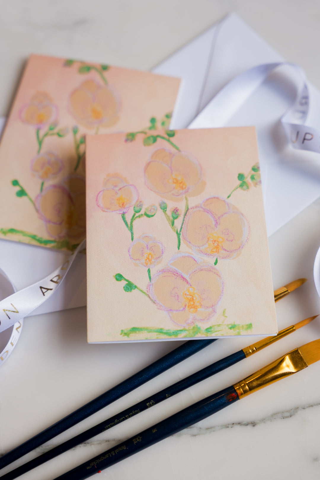 Card with Pink and yellow Orchids  and green leaves painted on it from Arden Upton's Flowers That Last Series sitting on a marble surface with paintbrushes. These Cards come in sets of 6 and make great gift ideas