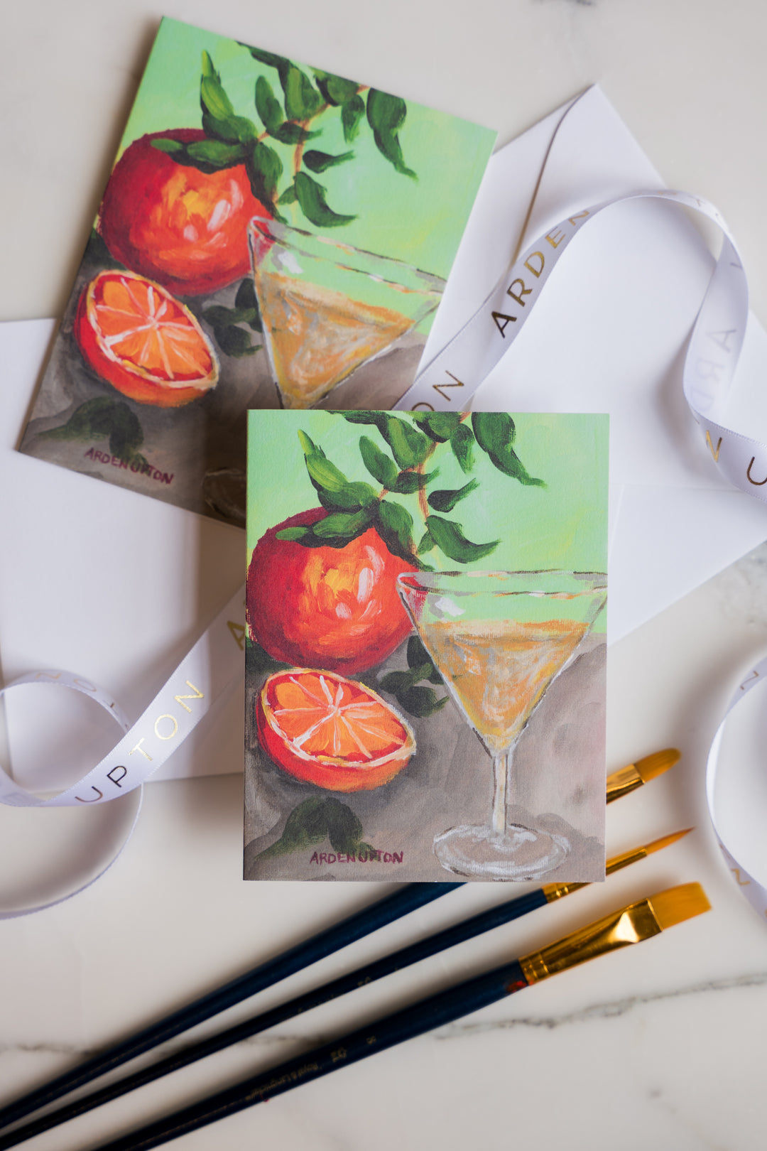 Two notecards with a beautiful, artistic original painting of oranges and a martini glass. The crds are sitting on a marble surface with art supplies. Print is from Arden Upton's Franks Table Series and the notecard set of 6 is a great gift idea