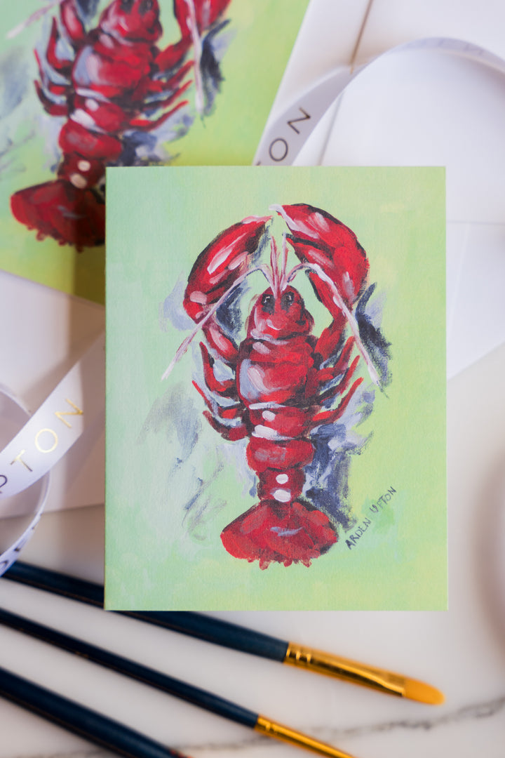 A photo of note cards from a set featuring Arden Upton's original lobster painting of a bright red, whimsical lobster on a green background. The note cards come in sets of 6 and are a great gift idea. In the photo, the note cards are surrounded by paintbrushes. 