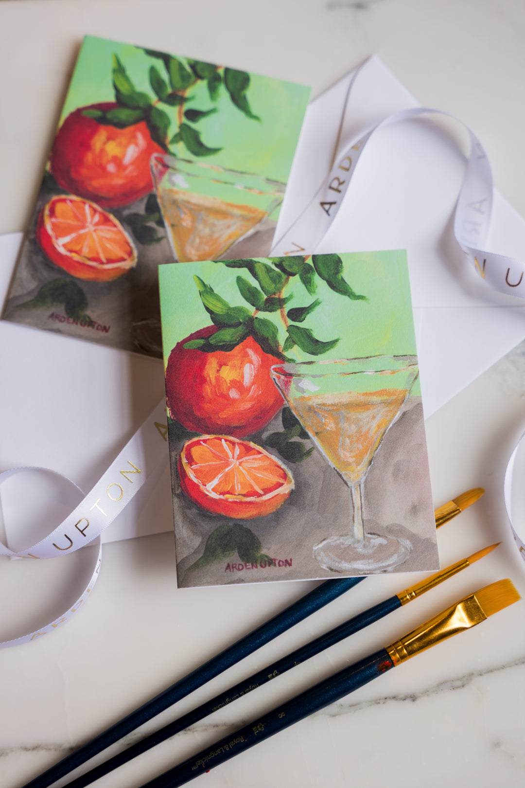 Two notecards with a beautiful, artistic original painting of oranges and a martini glass. The crds are sitting on a marble surface with art supplies. Print is from Arden Upton's Franks Table Series and the notecard set of 6 is a great gift idea