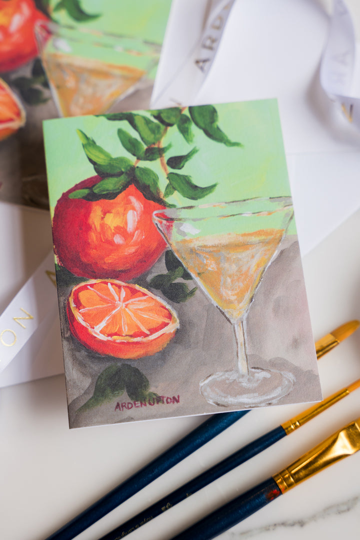 A folded notecard with a beautiful, artistic original painting of oranges and a martini glass. The crds are sitting on a marble surface with art supplies. Print is from Arden Upton's Franks Table Series and the notecard set of 6 is a great gift idea