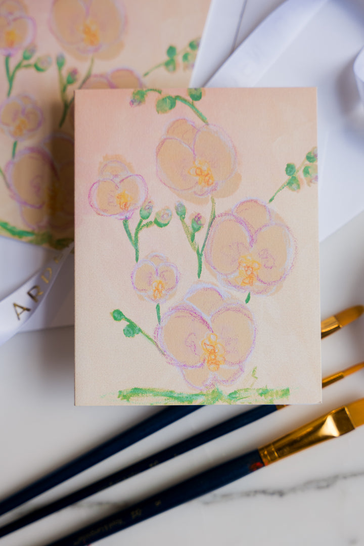 Card with Pink and yellow Orchids  and green leaves painted on it from Arden Upton's Flowers That Last Series sitting on a marble surface with paintbrushes. These Cards come in sets of 6 and make great gift ideas