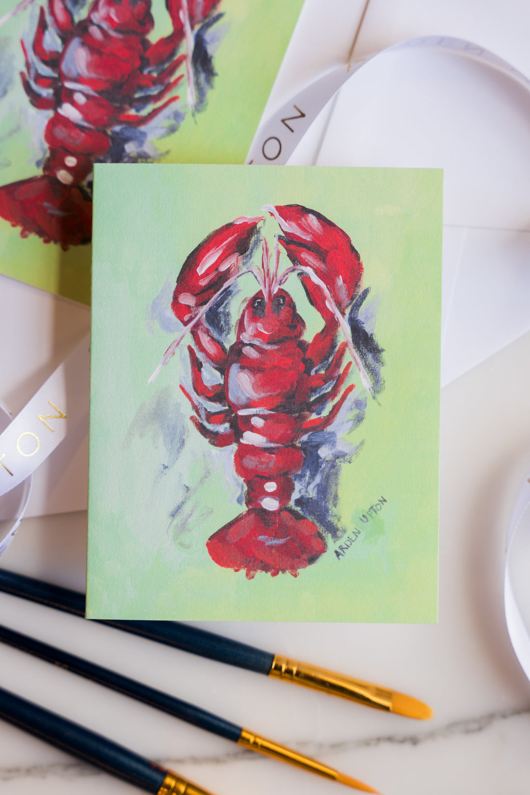  photo of a note card featuring Arden Upton's original lobster painting of a bright red, whimsical lobster on a green background. The note cards come in sets of 6 and are a great gift idea. In the photo, the note cards are surrounded by paintbrushes. 
