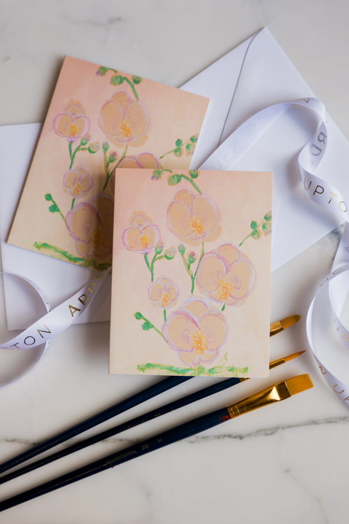 Card with Pink and yellow Orchids  and green leaves painted on it from Arden Upton's Flowers That Last Series sitting on a marble surface with paintbrushes. These Cards come in sets of 6 and make great gift ideas